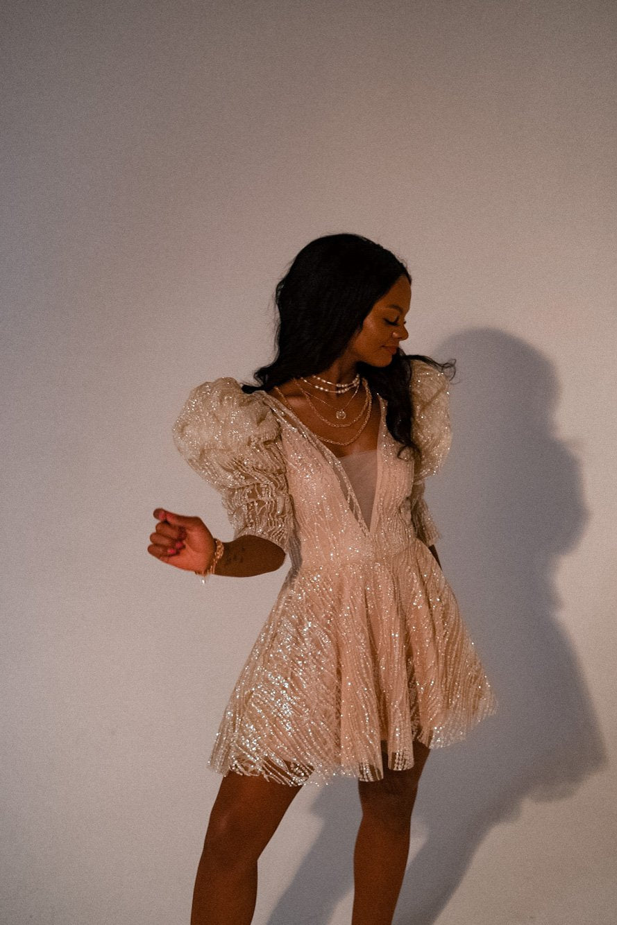 Champagne mini wedding dress twirled mid-step, flared glitter tulle skirt and ruffled sleeves against a plain wall, Soleil
