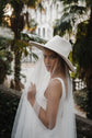 Side view of a raw-cut tulle wedding veil worn under a wide-brim hat, bride in a sleeveless gown among palms, Spell Veil