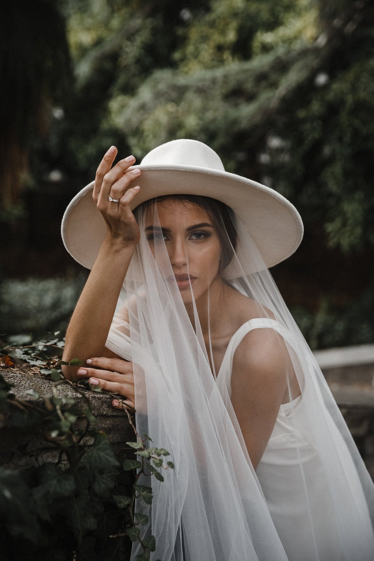 Fingertip wedding veil falling from under a wide-brim hat, bride leaning on an ivy-covered wall, shaded garden light, Spell Veil