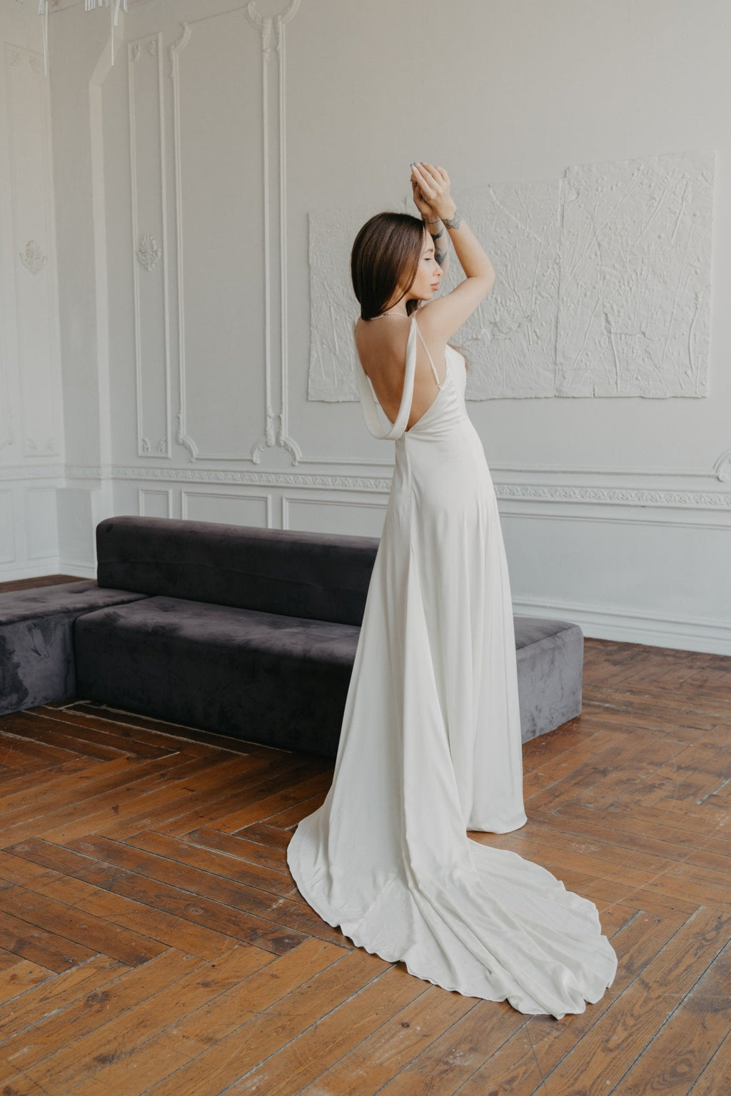 Model with arms raised in an open-back satin sample wedding dress, the long train curving across a wood floor, editorial, ZEPHYR Sample
