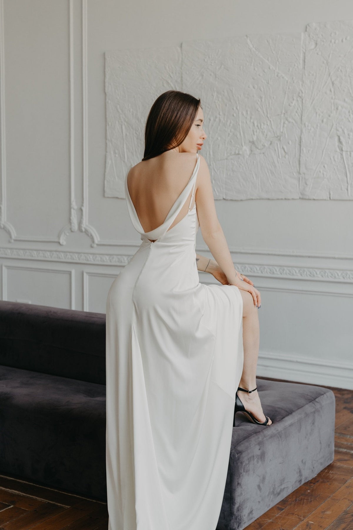 Back view of a bias satin sample wedding dress with a draped open back, model resting a foot on a bench, ZEPHYR Sample