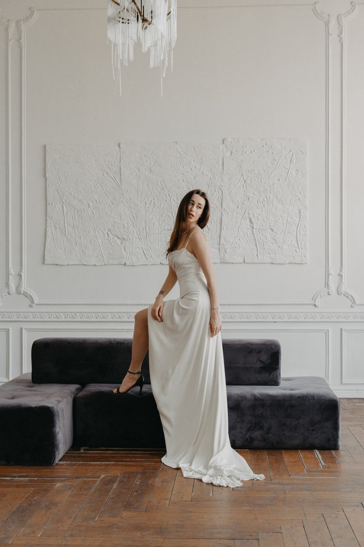 Model with one foot on a velvet bench in a slip sample wedding dress with a slit, panelled white room and chandelier, ZEPHYR Sample