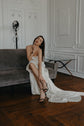 Model seated on a grey sofa in a satin sample wedding dress with a cowl neck and side slit, train spread on parquet, ZEPHYR Sample