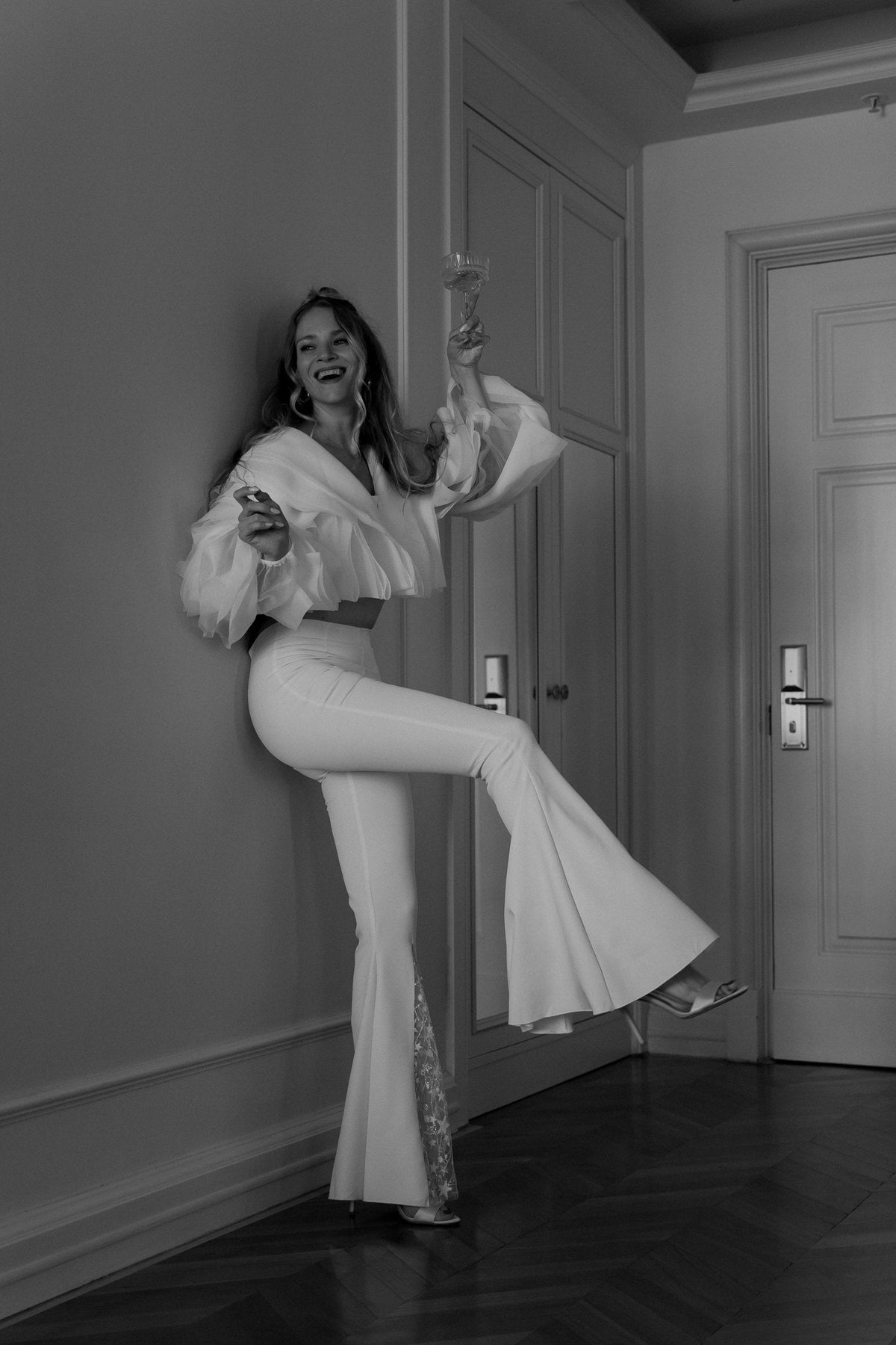 Black and white shot of a laughing bride kicking back a leg in flared bridal trousers against a hotel door, Sun Flares