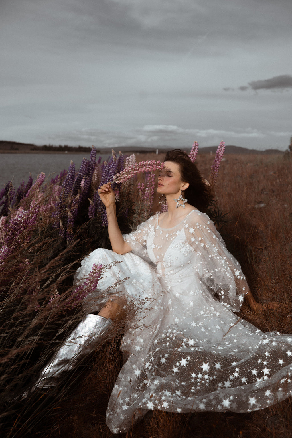 Bride lounging in a lupine wildflower field in silver boots, star lace wedding dress with sheer sleeves, Counting Stars Sample