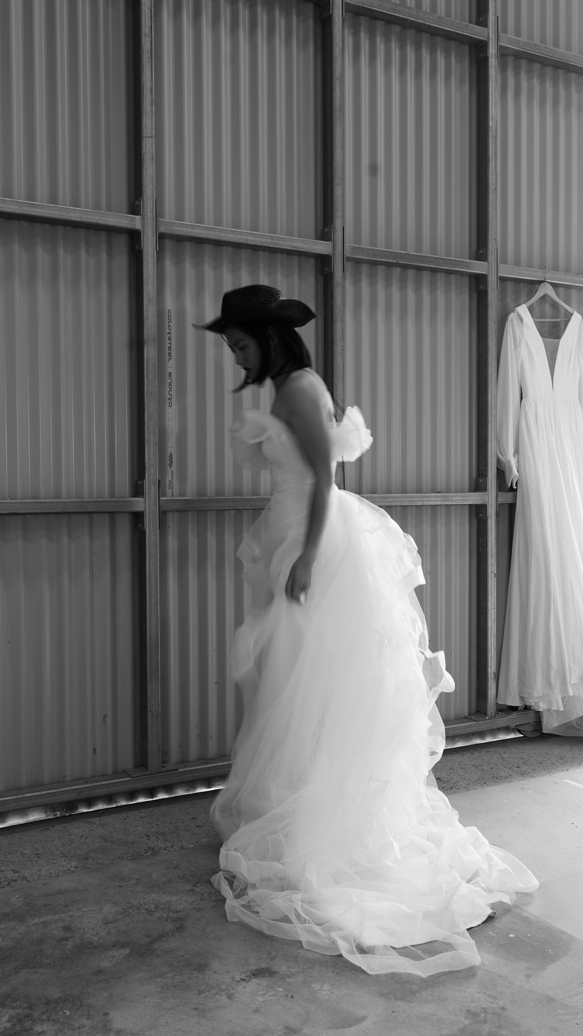 Bride in a black hat looking down in an off-shoulder wedding dress with a full tulle overskirt, warehouse, Terra