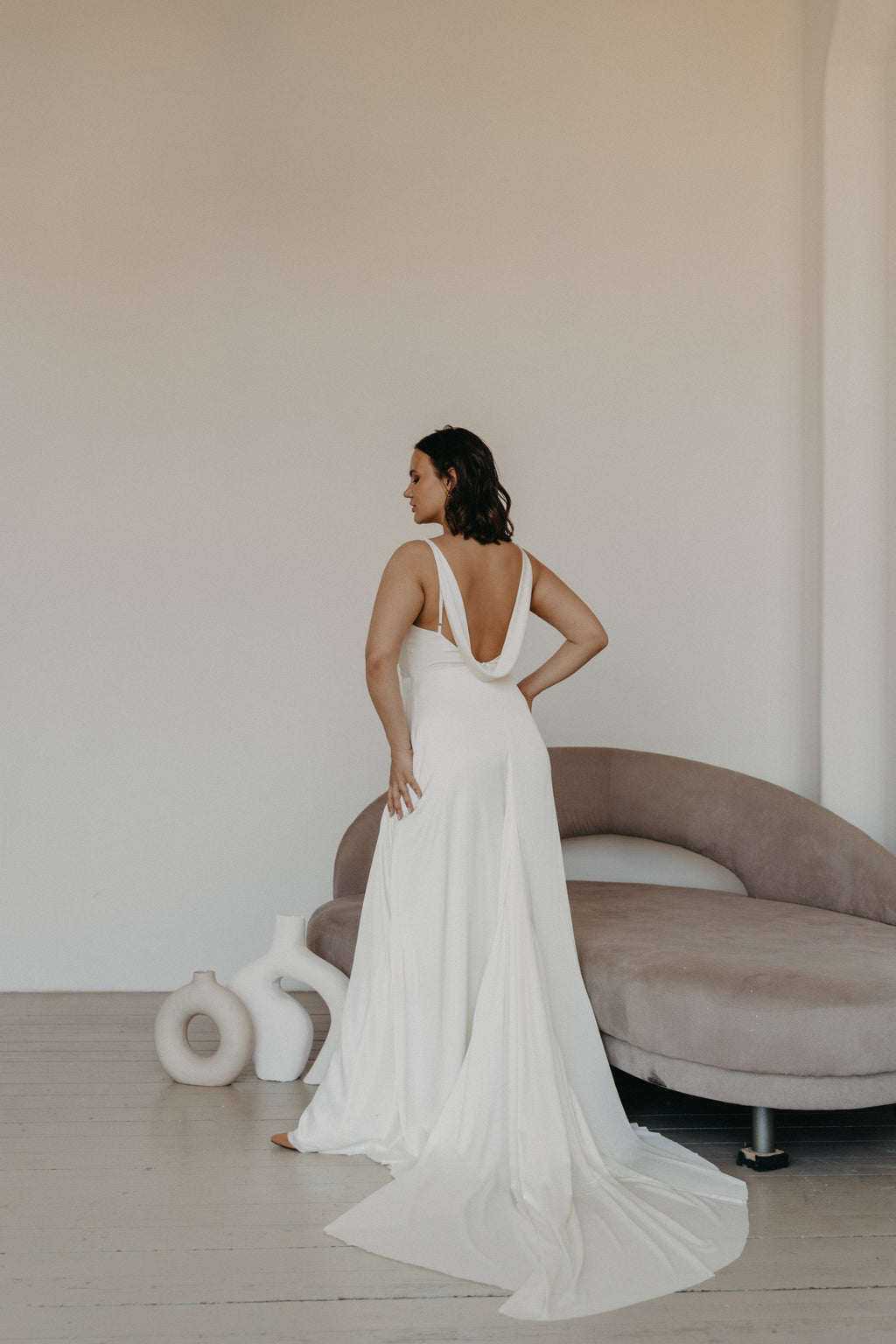 Satin sample wedding dress with thin straps and a draped cowl open back, model by a curved sofa, train on the floor, ZEPHYR Sample by Ephemeris
