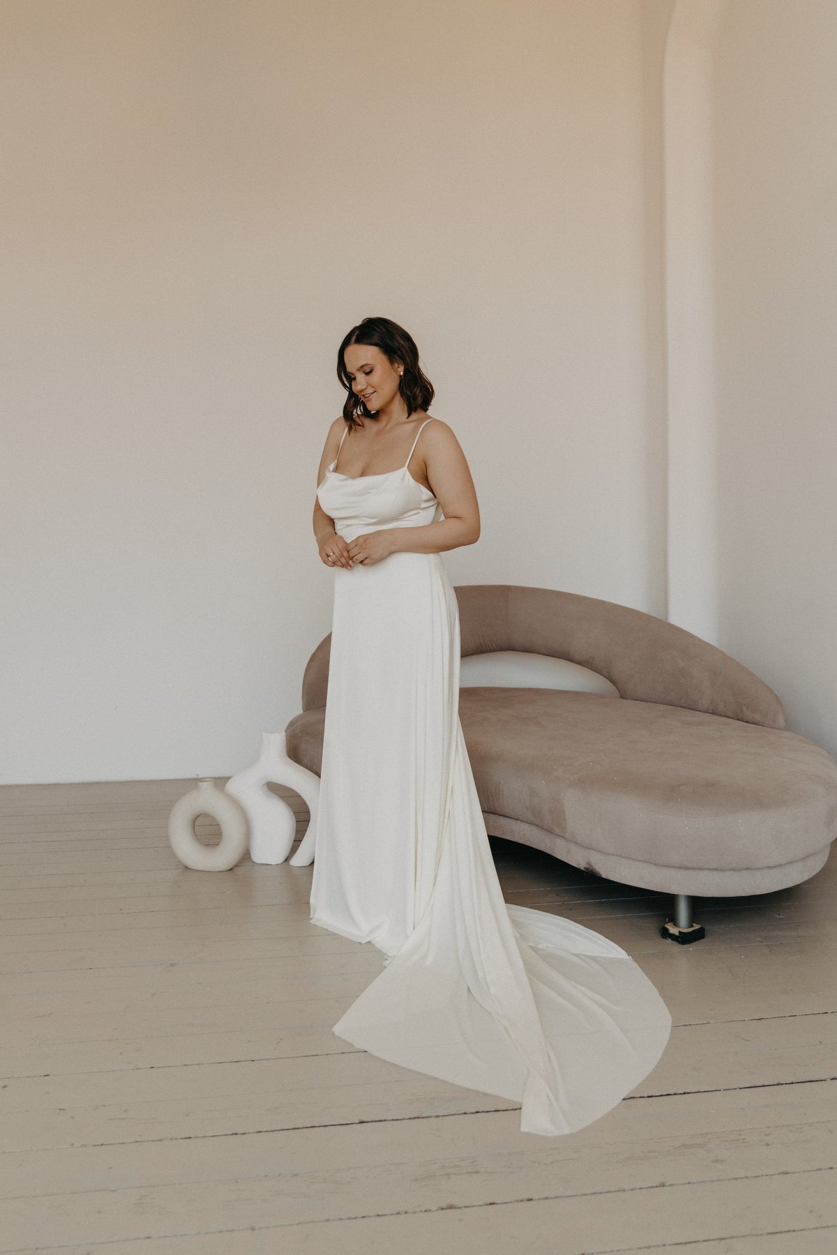 Model looking down in a satin slip sample wedding dress with thin straps, long train curling by a curved sofa, ZEPHYR Sample