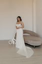 Model looking down in a satin slip sample wedding dress with thin straps, long train curling by a curved sofa, ZEPHYR Sample