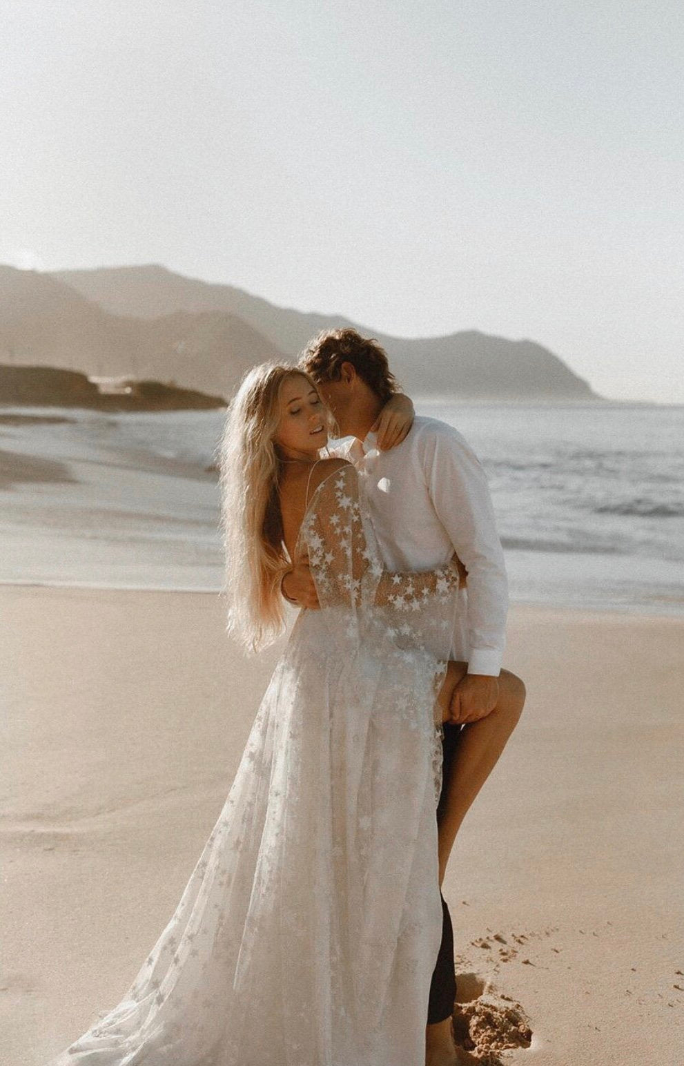Couple embracing on a beach at sunset, bride in a sheer star lace wedding dress with thin straps, Counting Stars