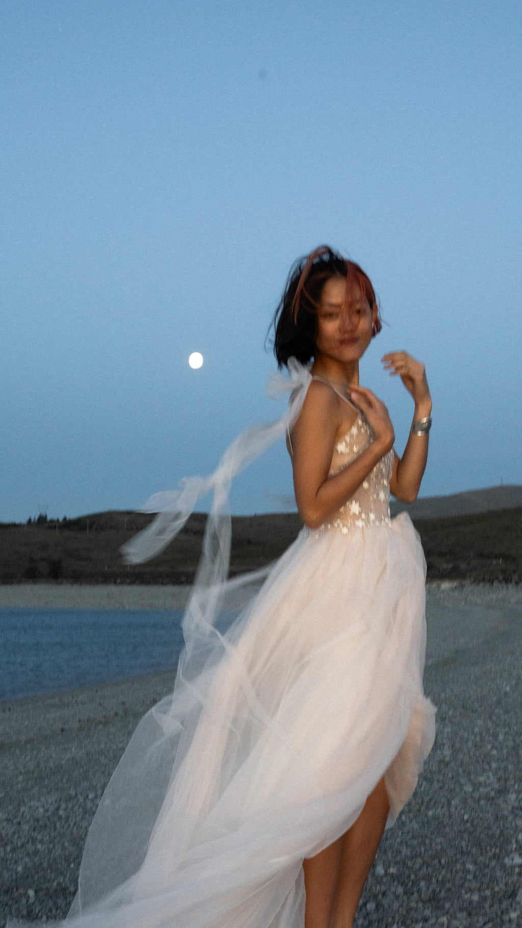 Bride posing by a lake under a rising moon, shoulder streamers of a plunge corset wedding dress blowing sideways, Make a Wish
