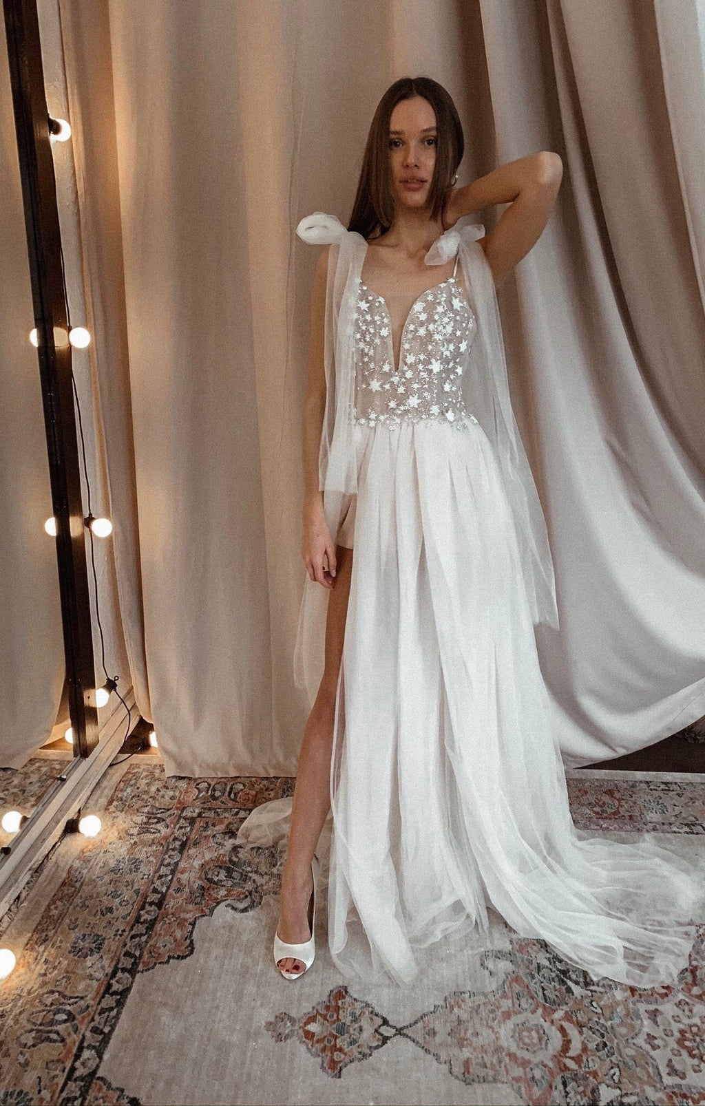 Bride with an arm raised under string lights, plunge star wedding dress with bow straps and a front slit, Make a Wish