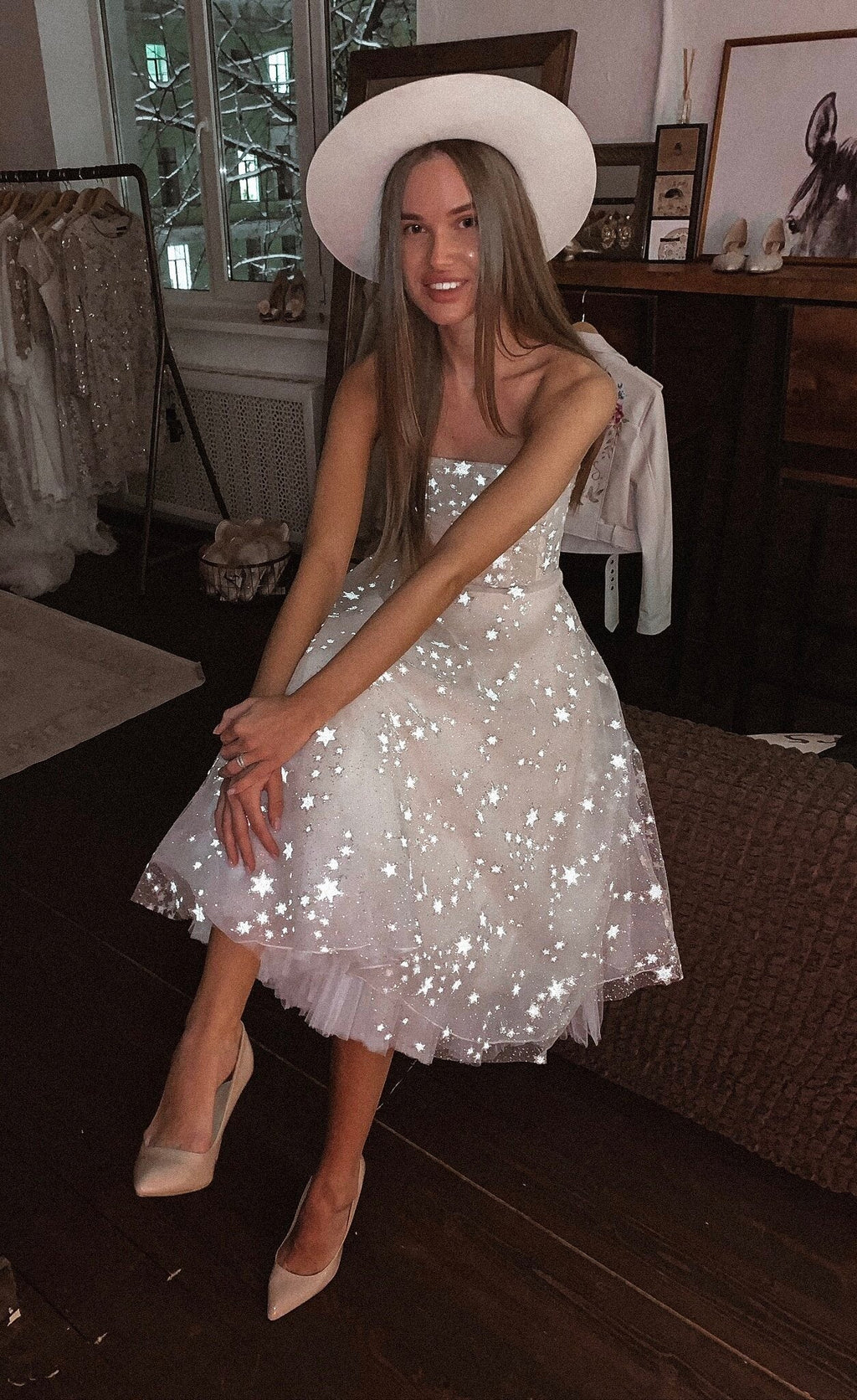 Seated model in a white hat, strapless star wedding dress with a midi tulle skirt flashing under the camera light, Wild Stars