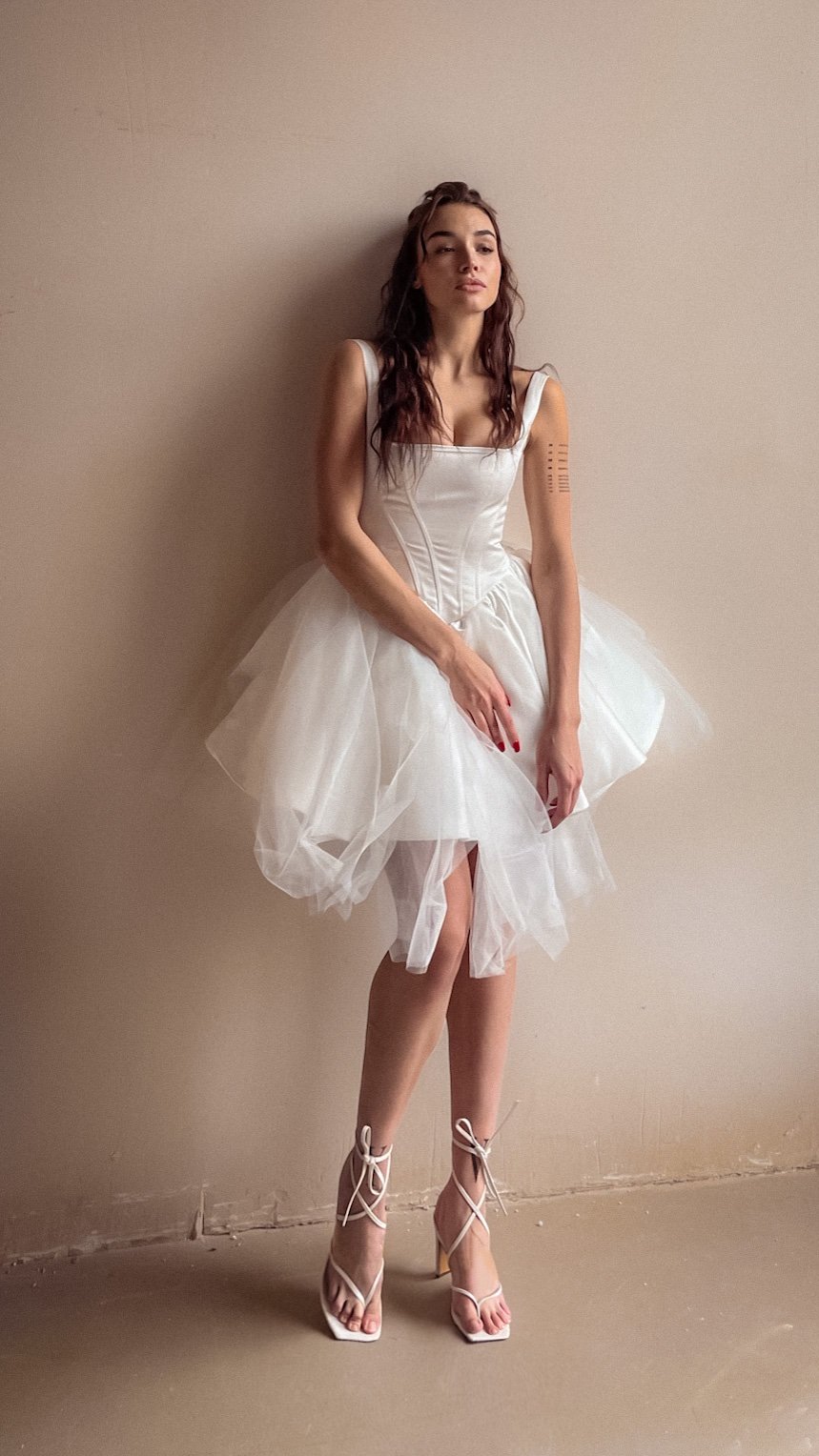 Model leaning on a plaster wall in a square-neck corset mini wedding dress, layered tulle skirt and lace-up sandals, Swan
