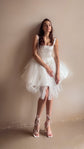 Model leaning on a plaster wall in a square-neck corset mini wedding dress, layered tulle skirt and lace-up sandals, Swan
