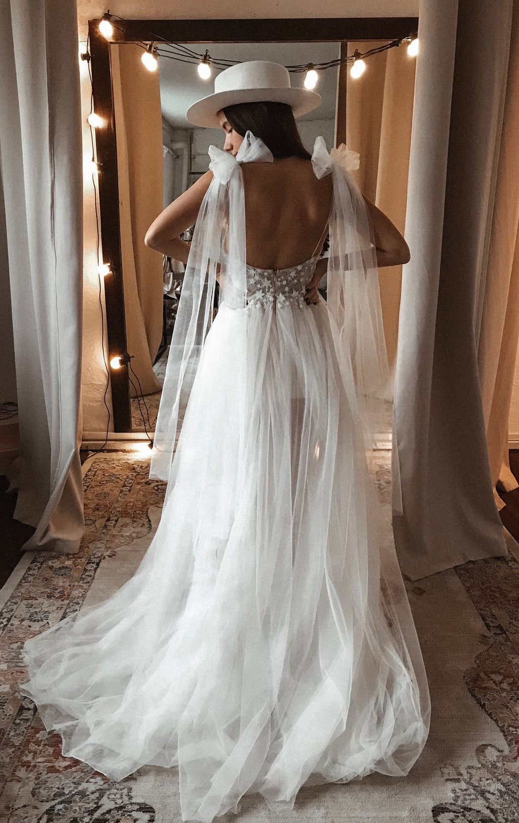 Back view of a low open-back star applique wedding dress with shoulder bows and a long skirt, string lights and hat, Make a Wish