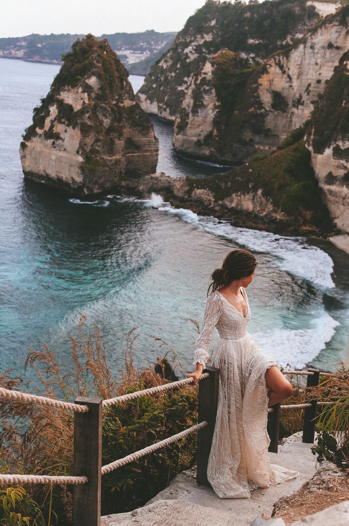 Bride on a cliff-top railing above a turquoise cove, long-sleeve ivory lace wedding dress with a side slit, Galaxy Ivory