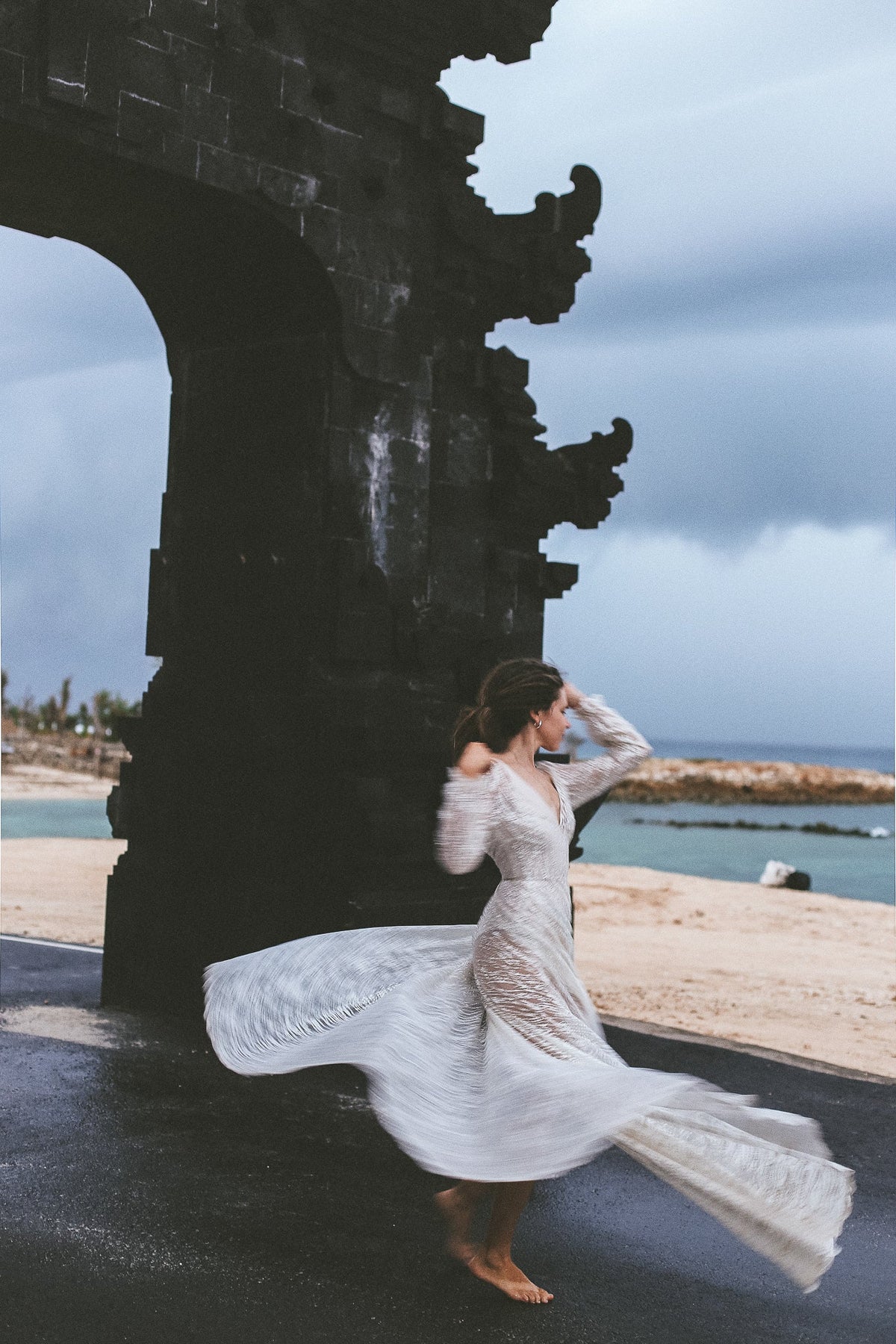 Barefoot bride spinning by a carved stone gate on the coast, ivory glitter lace wedding dress flaring in the wind, Galaxy Ivory