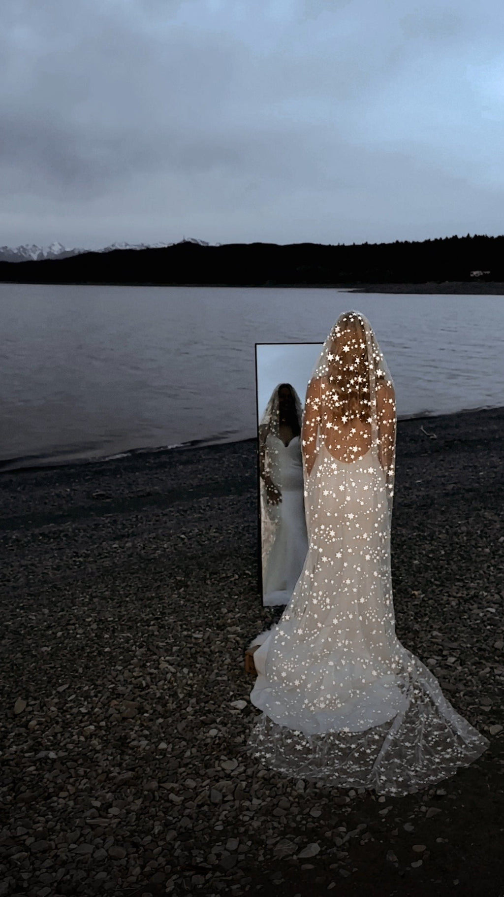 Cathedral star wedding veil on a pebble lakeshore at dusk, bride facing a standing mirror, back view, Milky Way Veil