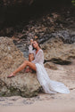 Bride perched on a beach boulder in an ivory glitter lace wedding dress with a front slit and train across the sand, Galaxy Ivory