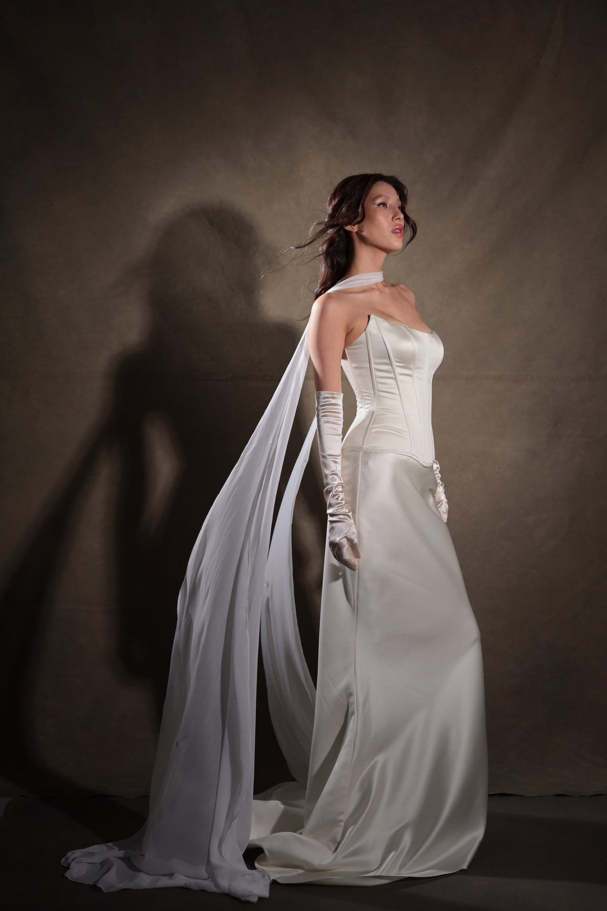 Bride in profile with a white bridal scarf at the neck flowing over her shoulder to a satin train, dark backdrop, Celeste Scarf