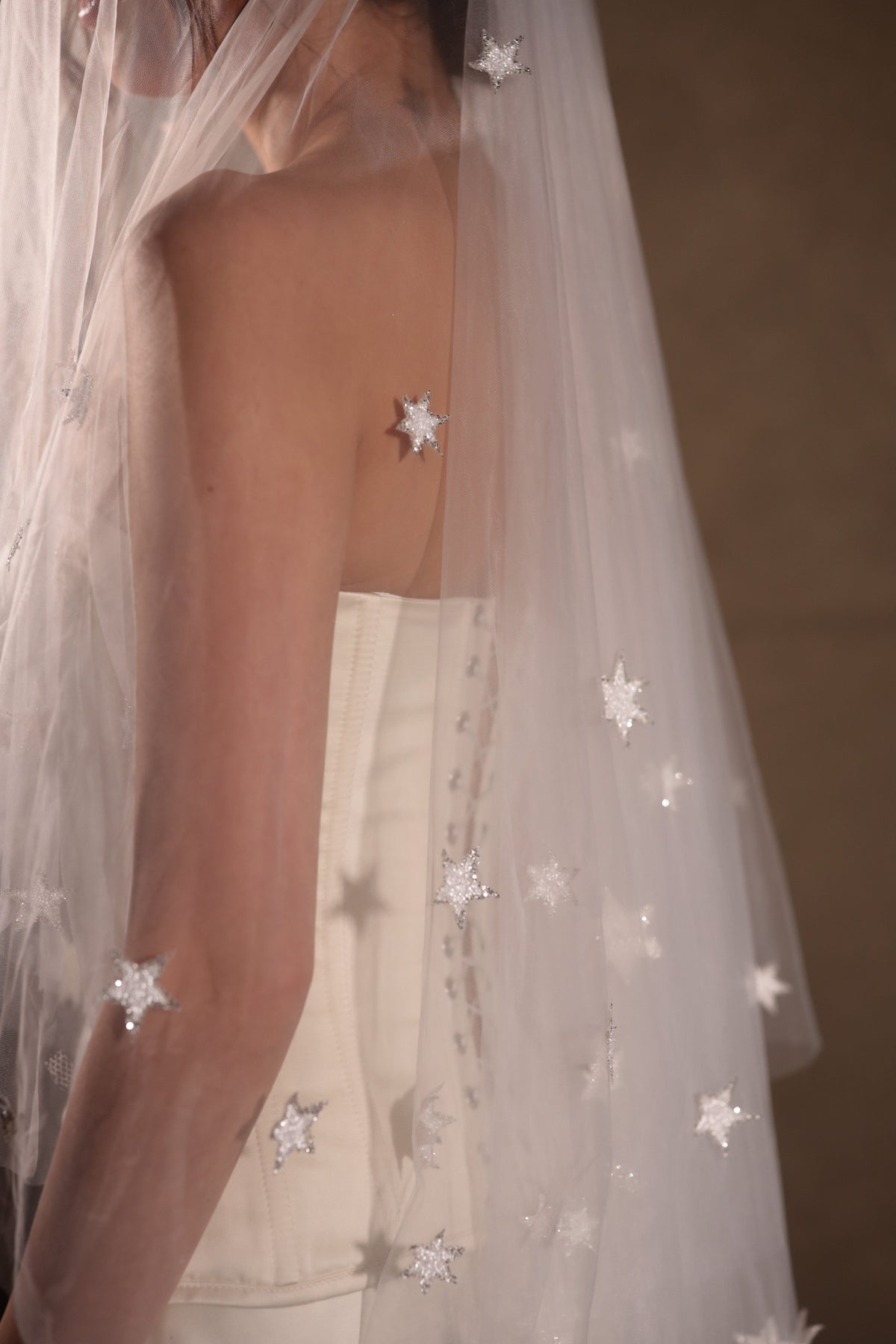 Close-up of hand-placed reflective stars on a sheer tulle wedding veil over a bare shoulder and corset back, Celestial Glow Veil