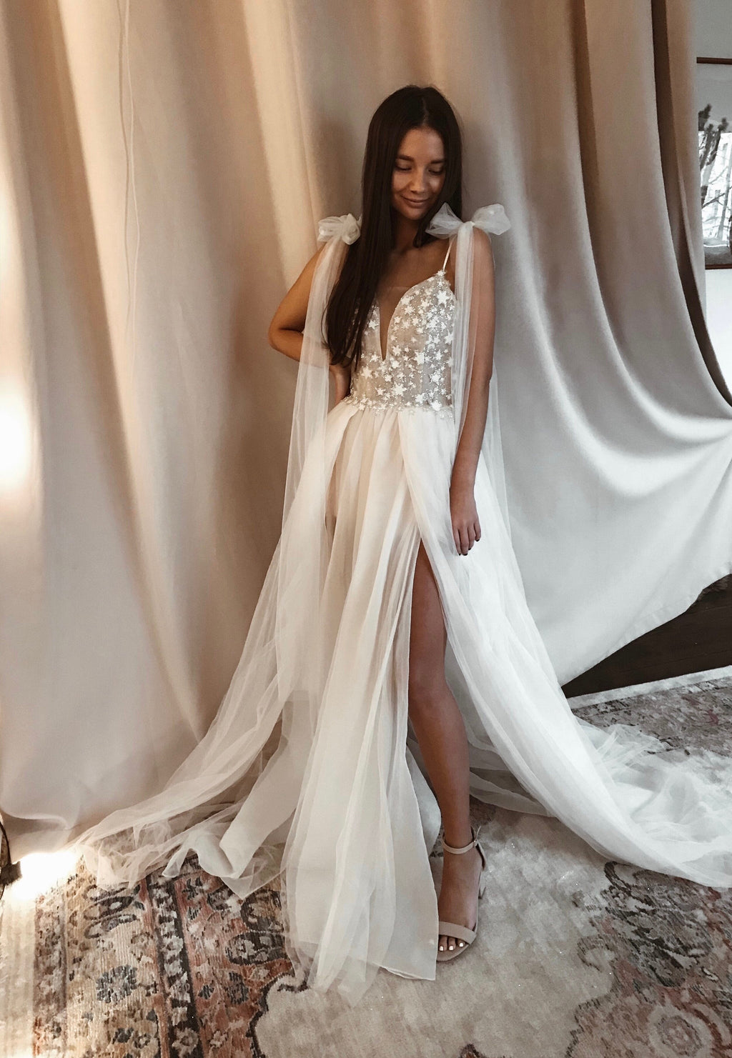 Star applique plunge wedding dress with bow ties at the shoulders and a slit skirt, bride on a rug by draped curtains, Make a Wish
