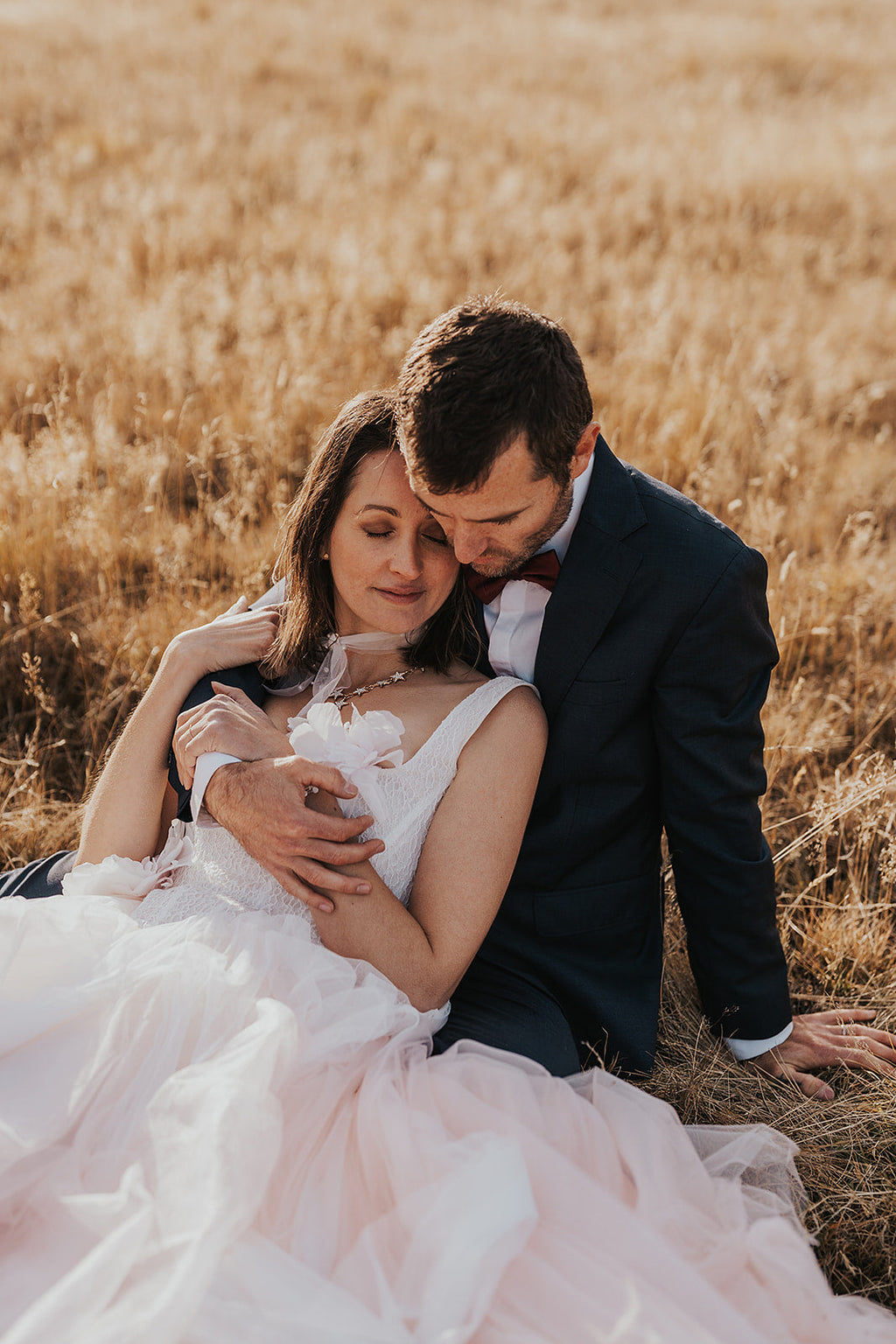 Elopement couple sitting in golden grass, bride in a blush wedding dress with lace corset bodice resting against the groom, Chrysanthemum