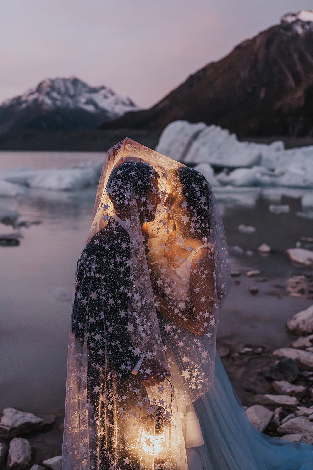 Couple embracing under a star wedding veil beside icebergs on a mountain lake at dusk, lantern in hand, Milky Way Veil