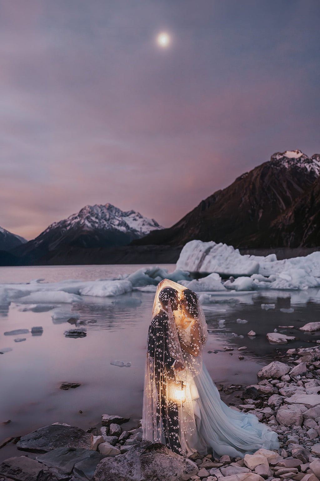 Elopement couple sheltering under a star wedding veil with a lantern on a glacier lake shore, snowy peaks and moon, Milky Way Veil