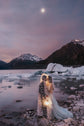 Elopement couple sheltering under a star wedding veil with a lantern on a glacier lake shore, snowy peaks and moon, Milky Way Veil