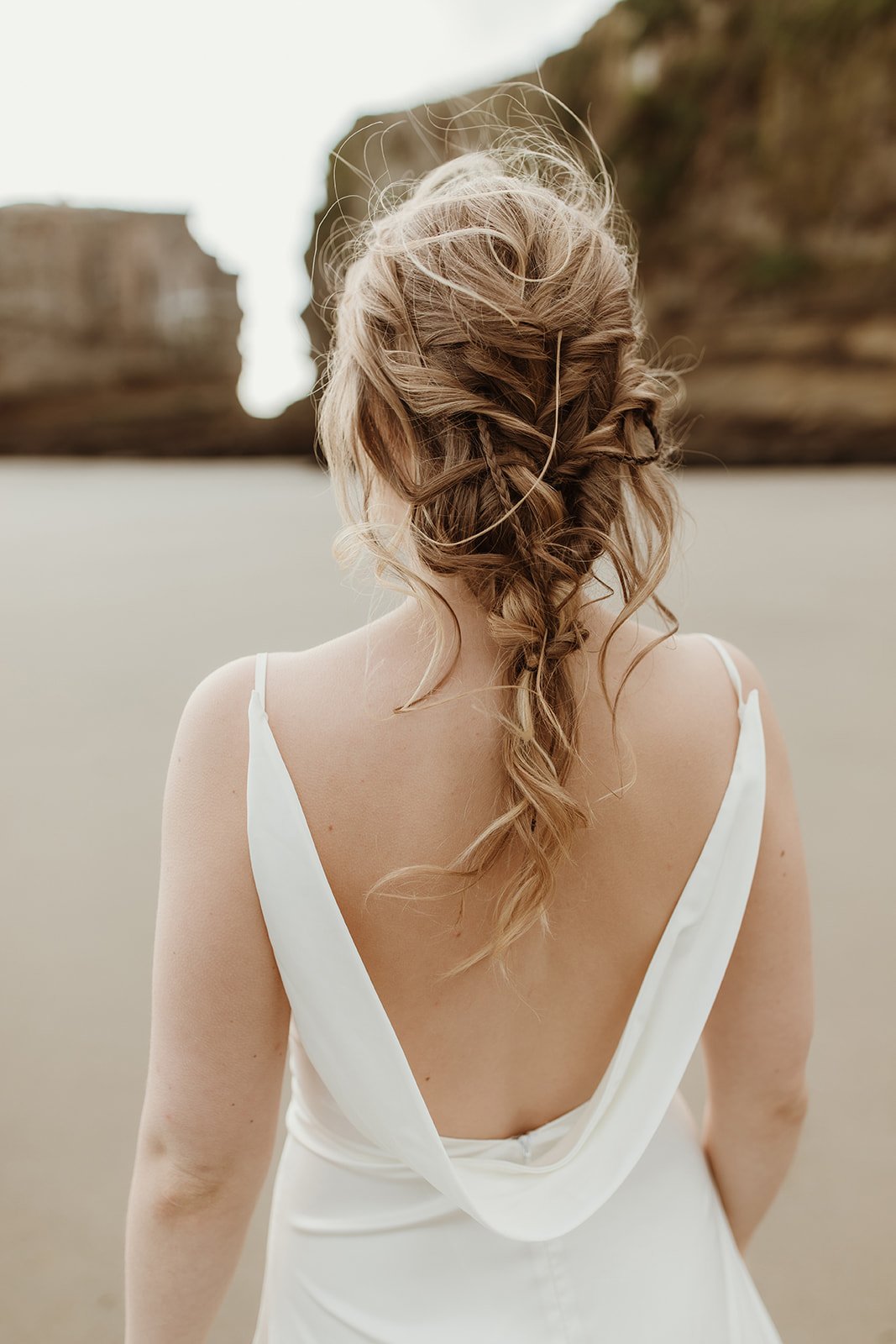 Close back view of a satin sample wedding dress with a draped cowl back and loose braid, beach blurred behind, ZEPHYR Sample