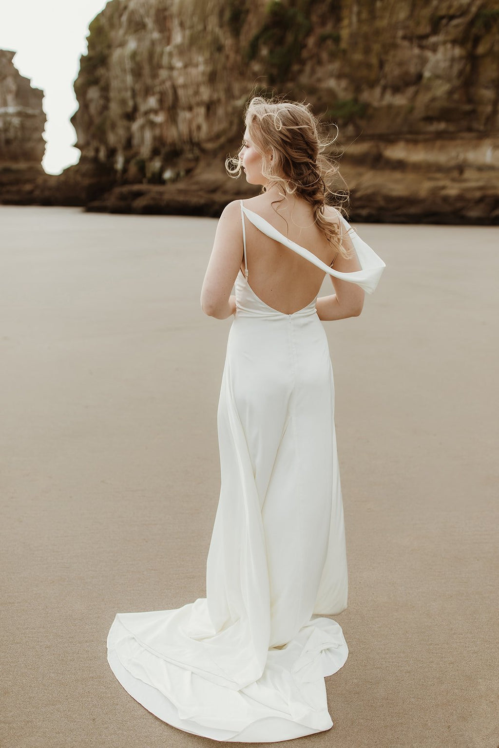 Back view of a bride on the sand in a satin sample wedding dress, one strap slipped and the cowl back open, train behind, ZEPHYR Sample