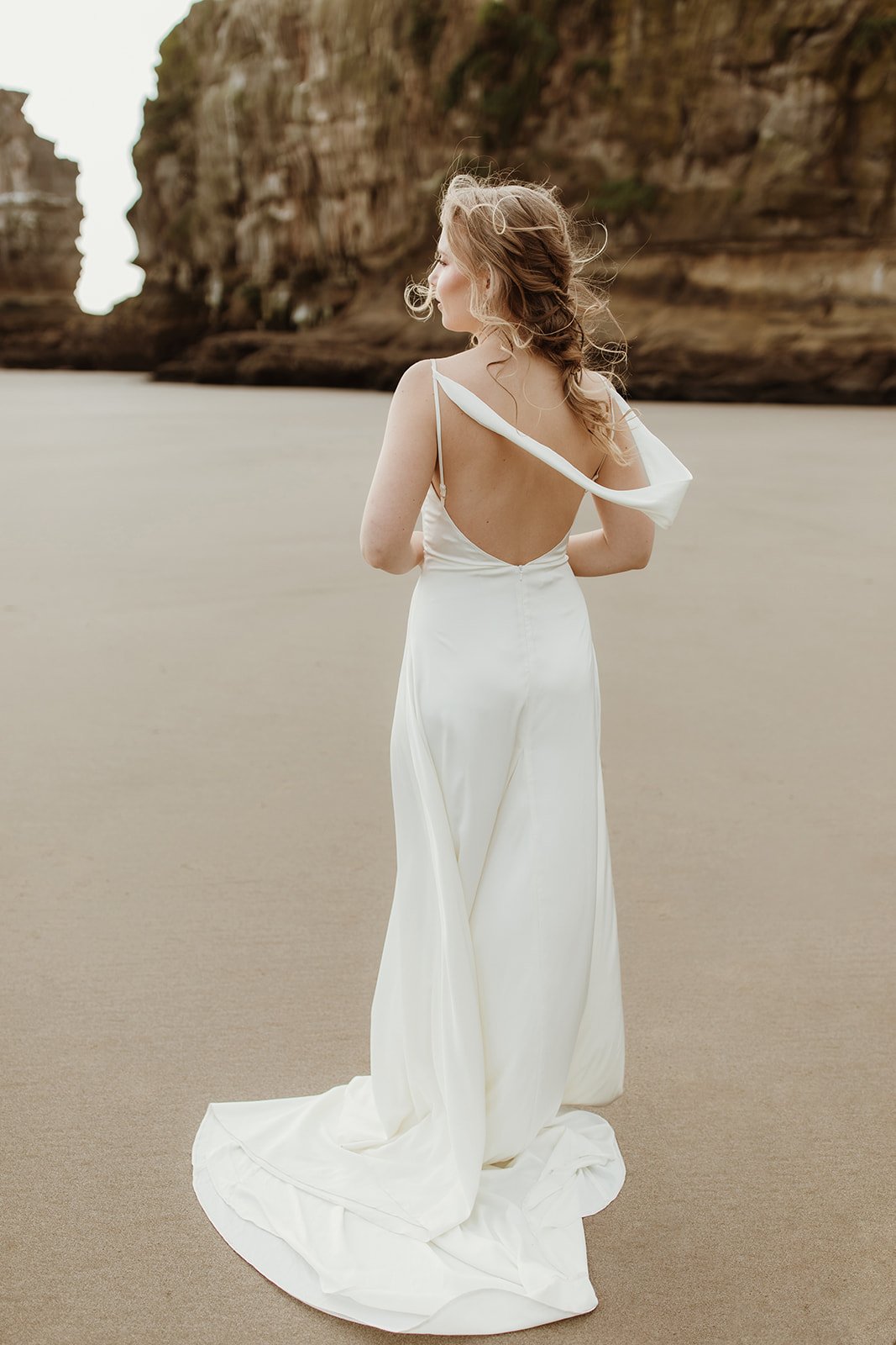 Back view of a bride on the sand in a satin sample wedding dress, one strap slipped and the cowl back open, train behind, ZEPHYR Sample