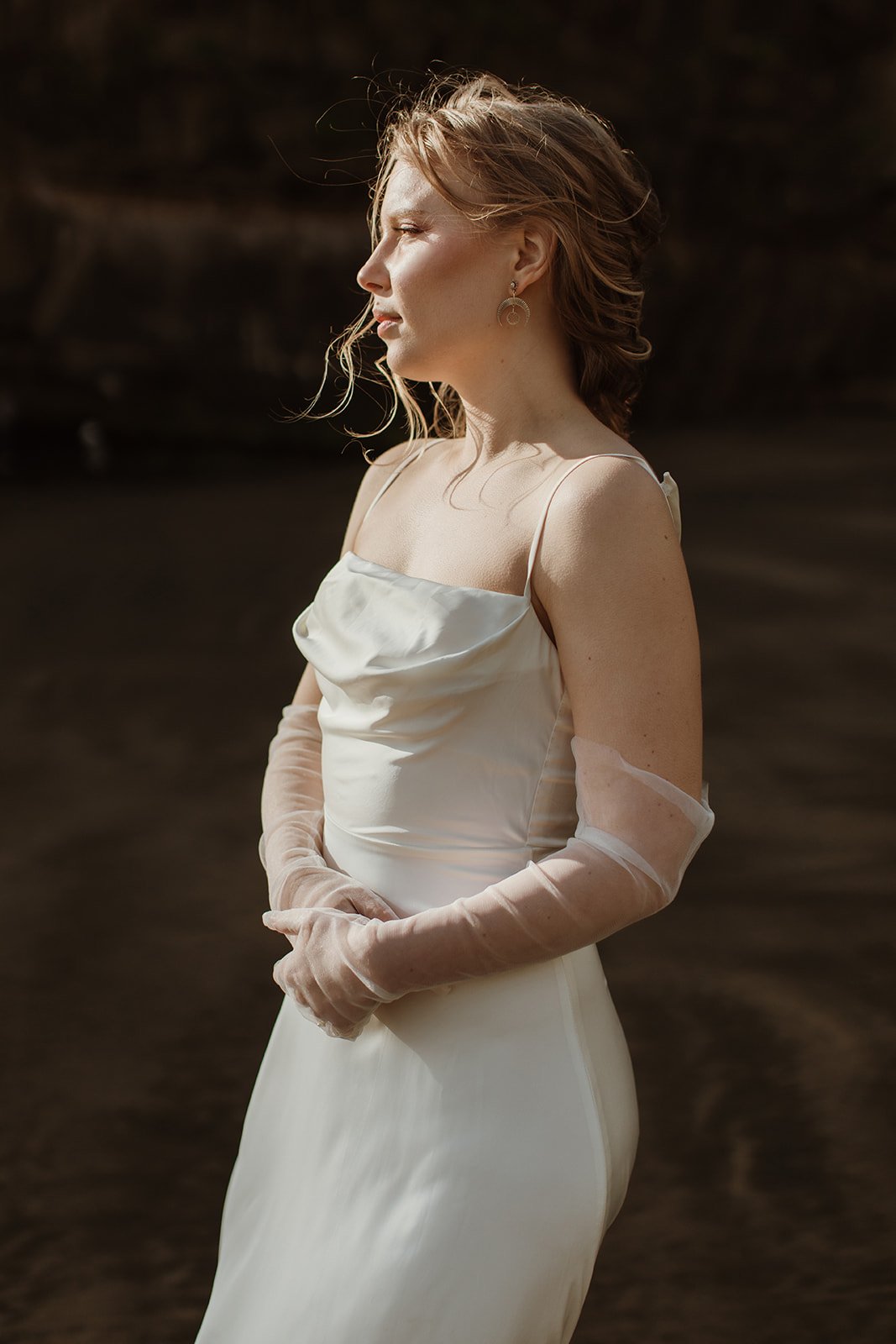 Profile of a bride in sheer gloves wearing a satin cowl neck sample wedding dress, dark cliff behind, soft light, ZEPHYR Sample