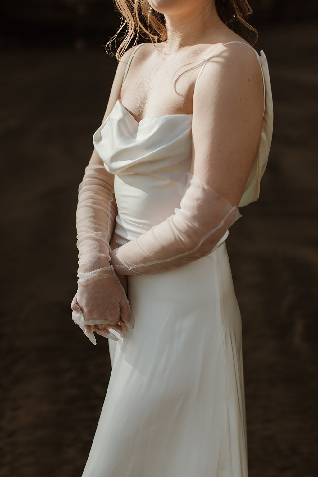 Crop of a satin sample wedding dress with a cowl neckline and spaghetti straps, hands clasped in sheer gloves, ZEPHYR Sample