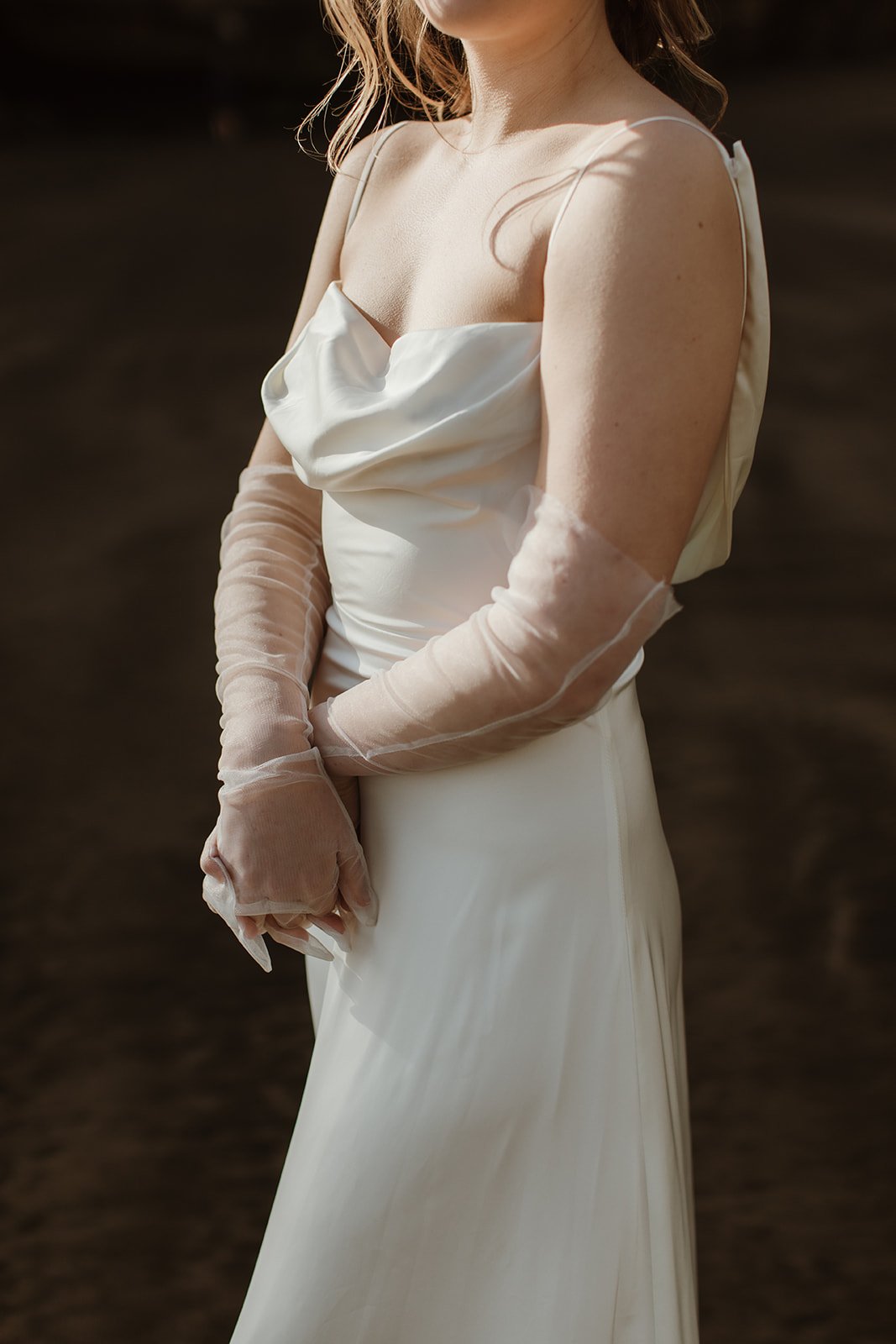 Crop of a satin sample wedding dress with a cowl neckline and spaghetti straps, hands clasped in sheer gloves, ZEPHYR Sample