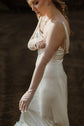 Close side view of a satin slip sample wedding dress with a cowl neckline, model pulling on sheer gloves against a dark backdrop, ZEPHYR Sample
