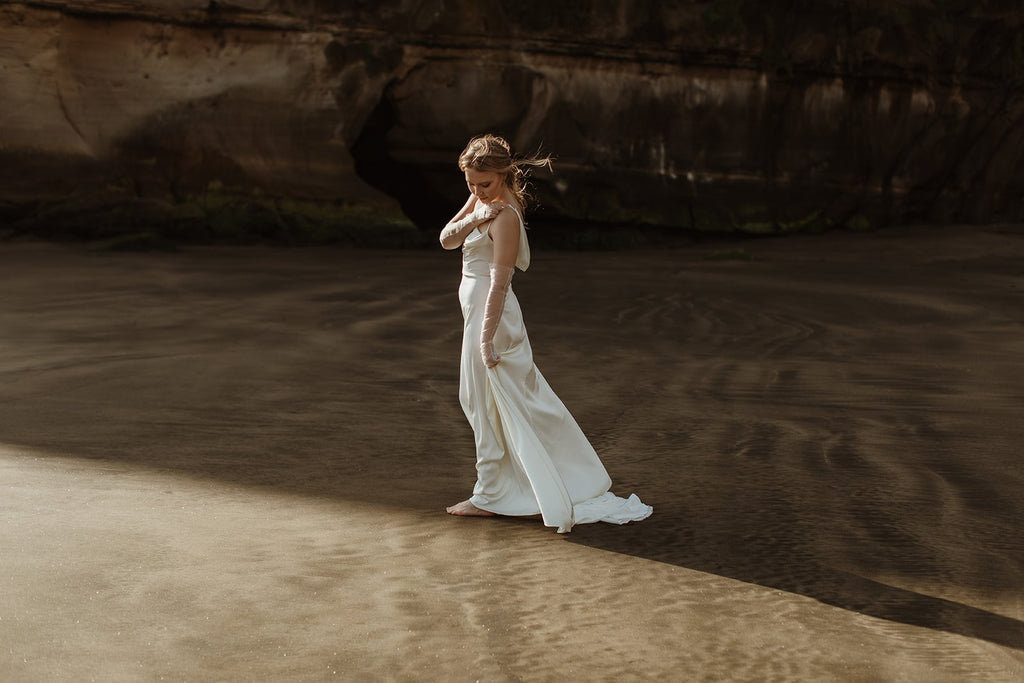 Wide view of a bride on a beach below cliffs in a satin slip sample wedding dress, train trailing on wet sand, elopement, ZEPHYR Sample