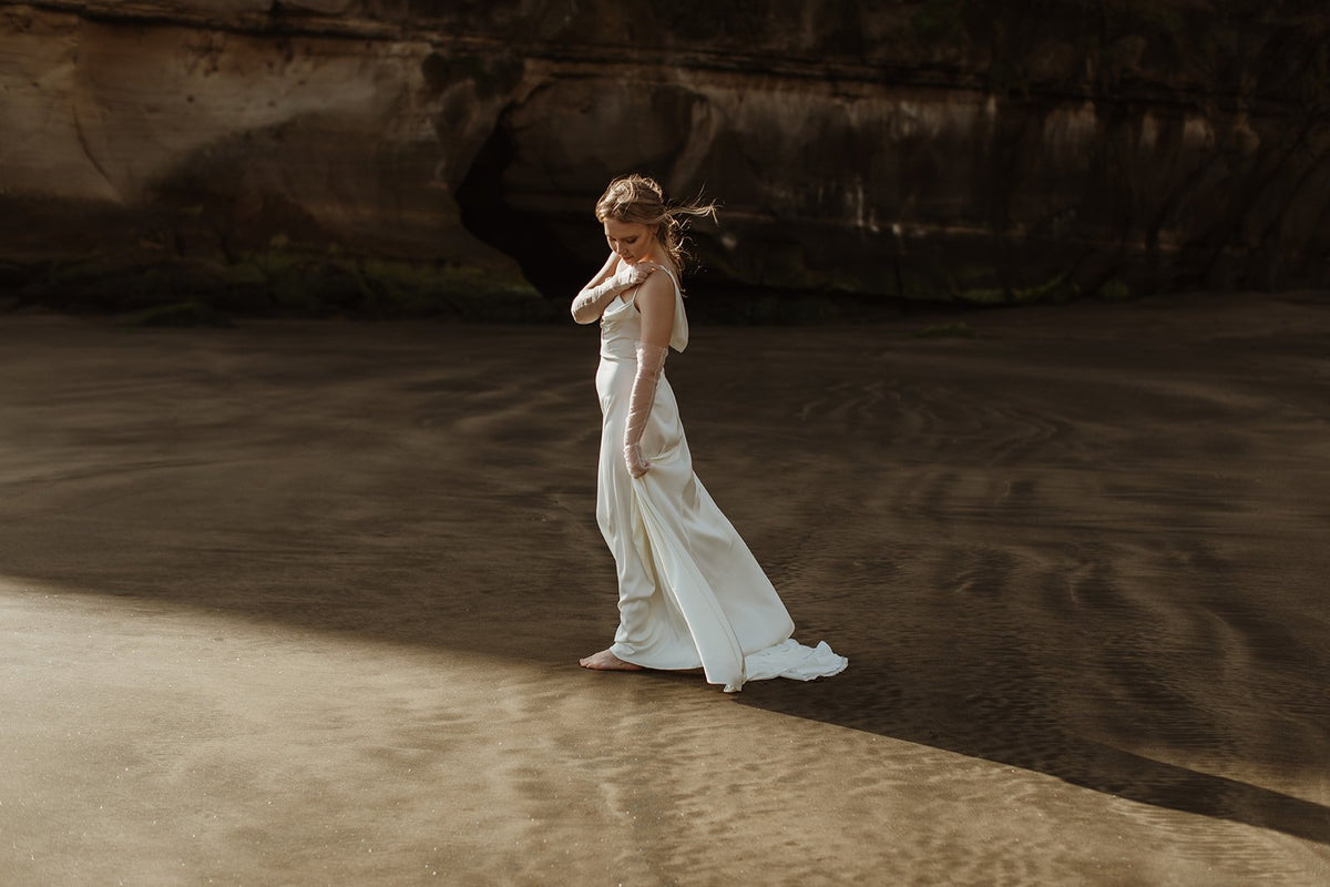 Wide view of a bride on a beach below cliffs in a satin slip sample wedding dress, train trailing on wet sand, elopement, ZEPHYR Sample