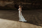 Wide view of a bride on a beach below cliffs in a satin slip sample wedding dress, train trailing on wet sand, elopement, ZEPHYR Sample