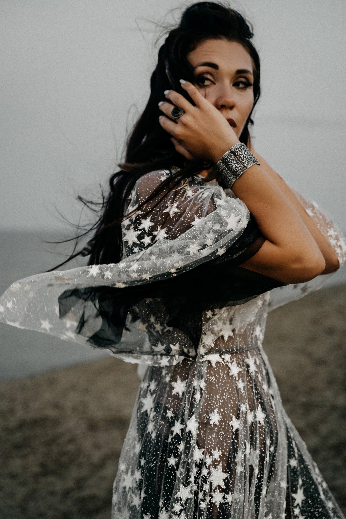 Close-up of sheer ivory star lace sleeves over a black tulle wedding dress, bride touching her face on a cliff, ASH Counting Stars