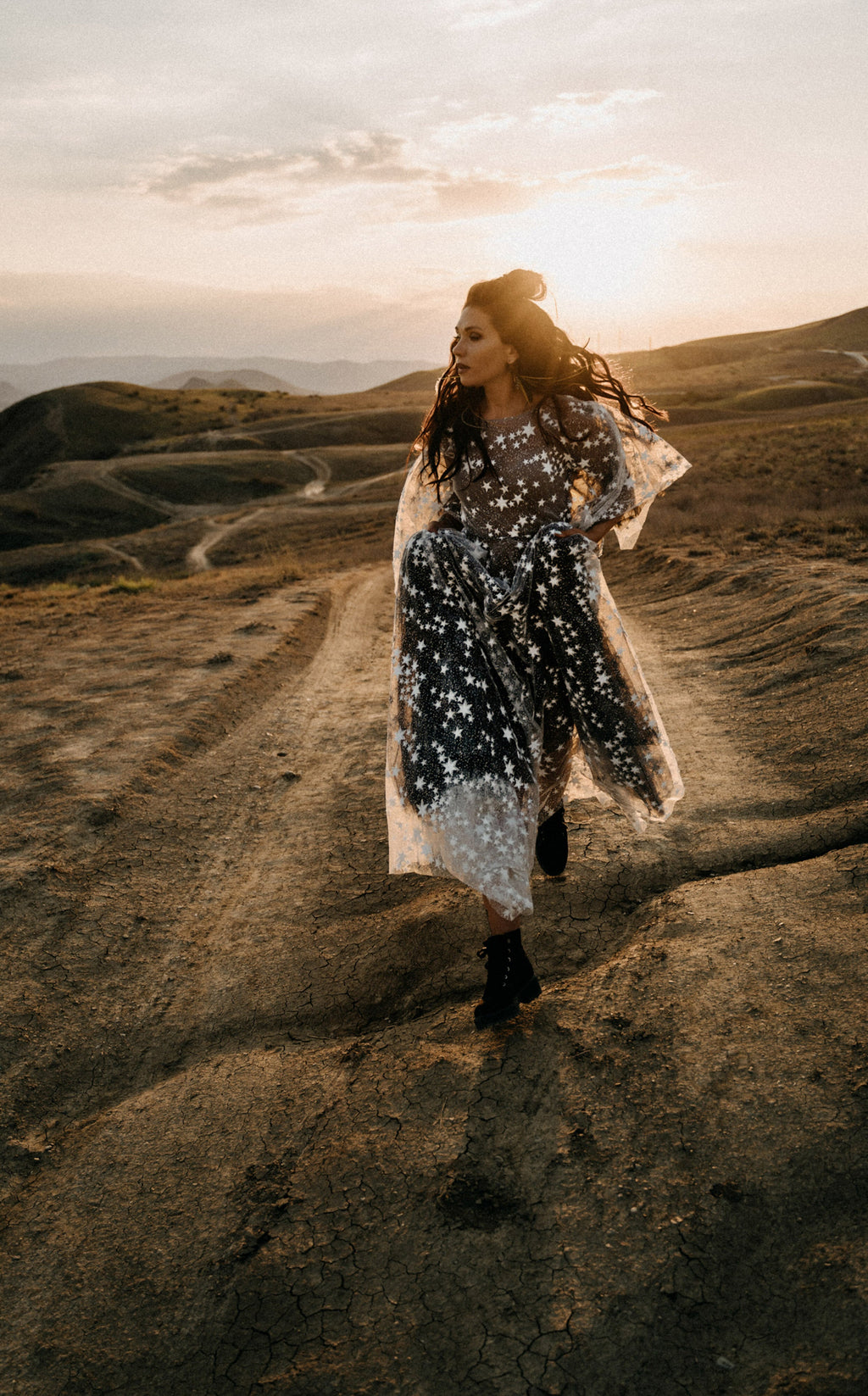 Full-length bride walking a dirt road in boots, black and ivory star lace wedding dress blowing at sunset, ASH Counting Stars