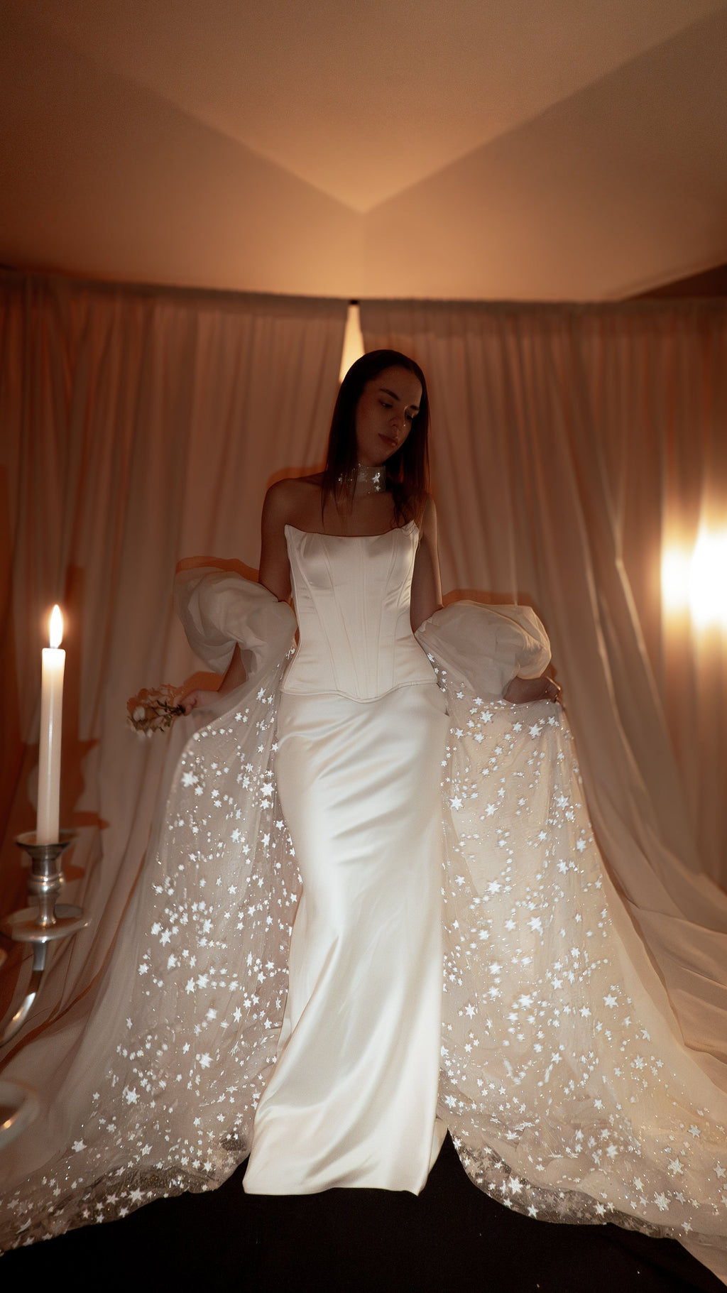 Ivory bridal cape with puff sleeves and a star-scattered train over a satin corset gown, candlelit room. Aura Cape by Ephemeris