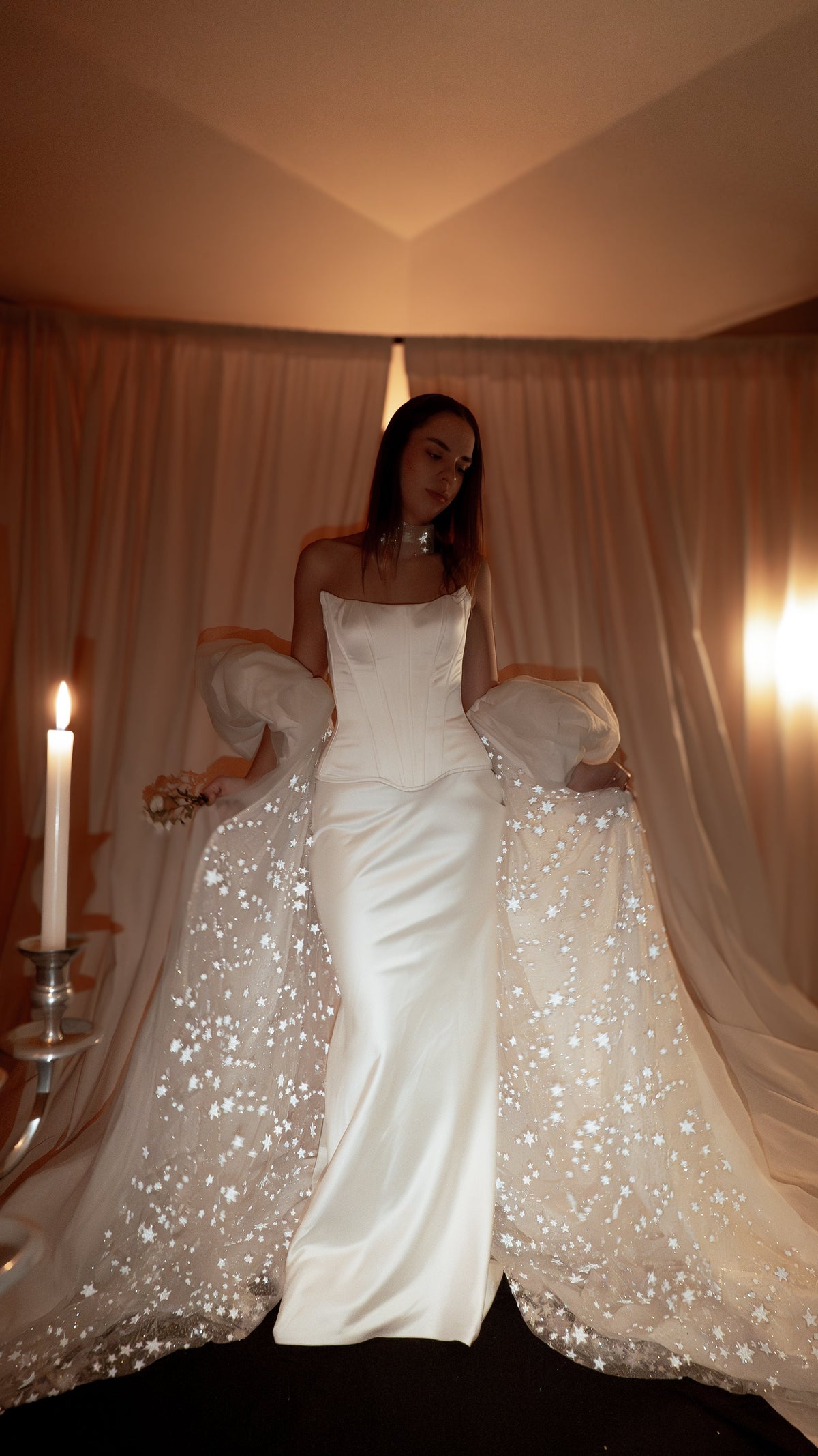 Ivory bridal cape with puff sleeves and a star-scattered train over a satin corset gown, candlelit room. Aura Cape by Ephemeris