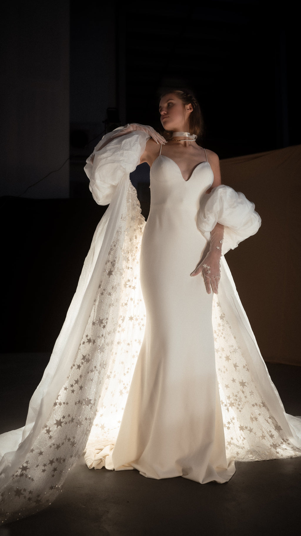 Bridal cape with a star-scattered train lit from behind in a dark studio, puff sleeves and an ivory slip gown, editorial pose, Aura Cape
