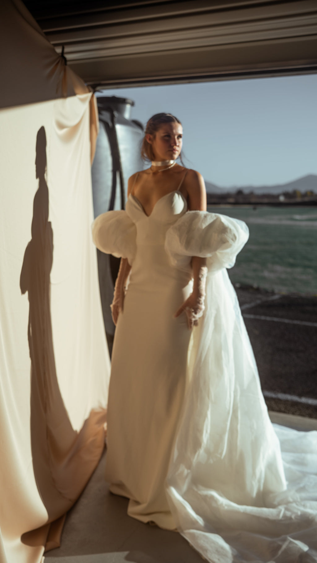 Bride in an ivory bridal cape and slip gown at an open garage door, chiffon train on the floor and her shadow on the wall, Aura Cape