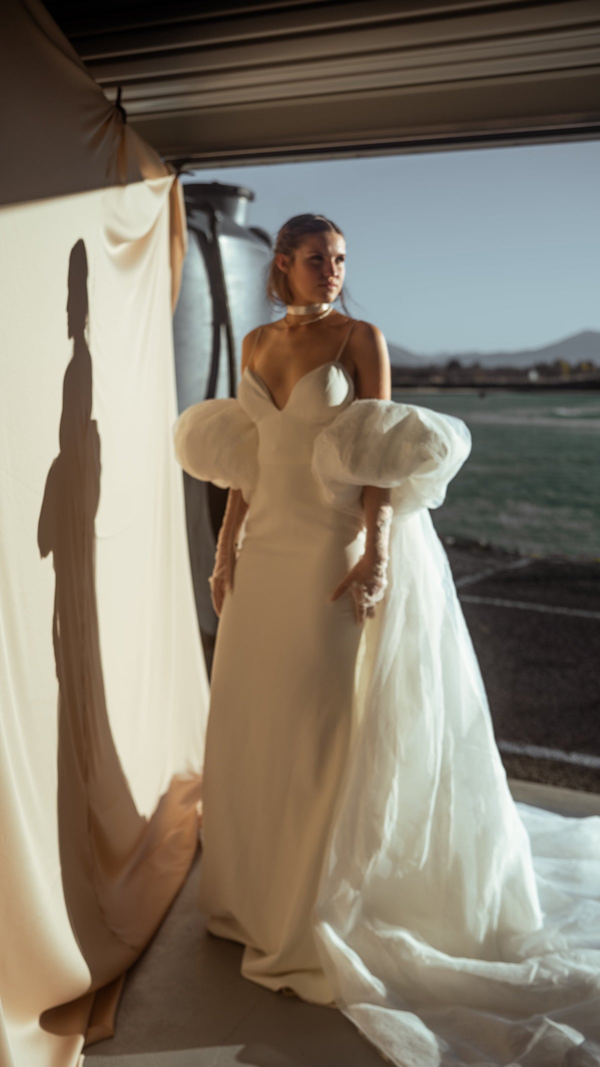 Bride in an ivory bridal cape and slip gown at an open garage door, chiffon train on the floor and her shadow on the wall, Aura Cape