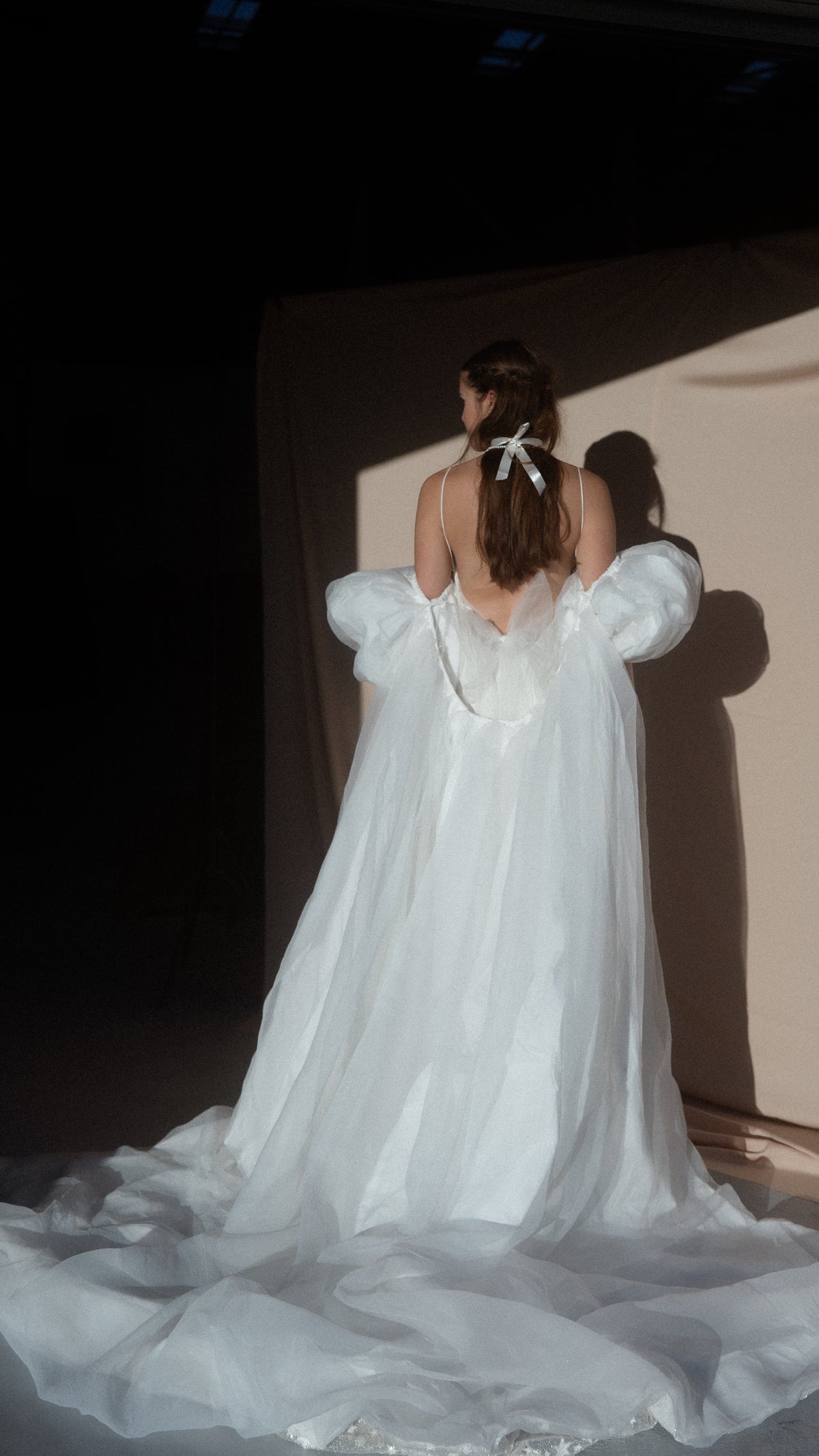 Back view of an ivory bridal cape with puff sleeves and a long chiffon train pooled on the floor, ribbon in the hair, Aura Cape