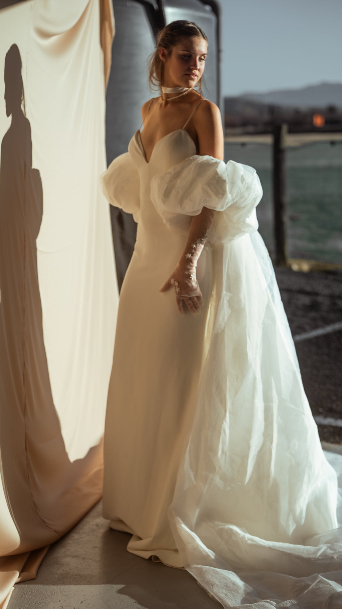 Bride turning at a doorway in an ivory chiffon bridal cape with puff sleeves, sheer gloves and a slip gown, soft daylight, Aura Cape
